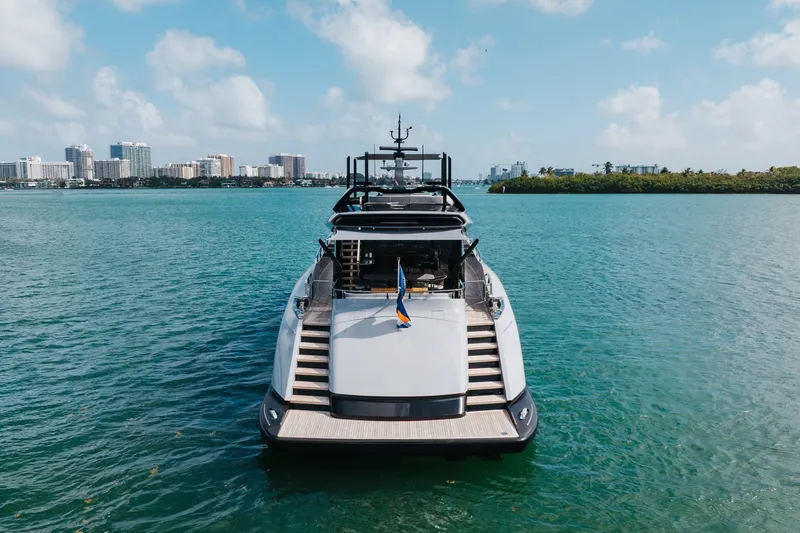 Slide: The Image of 2024 Mangusta 104 REV yacht on serene blue water with city skyline backdrop. - 7