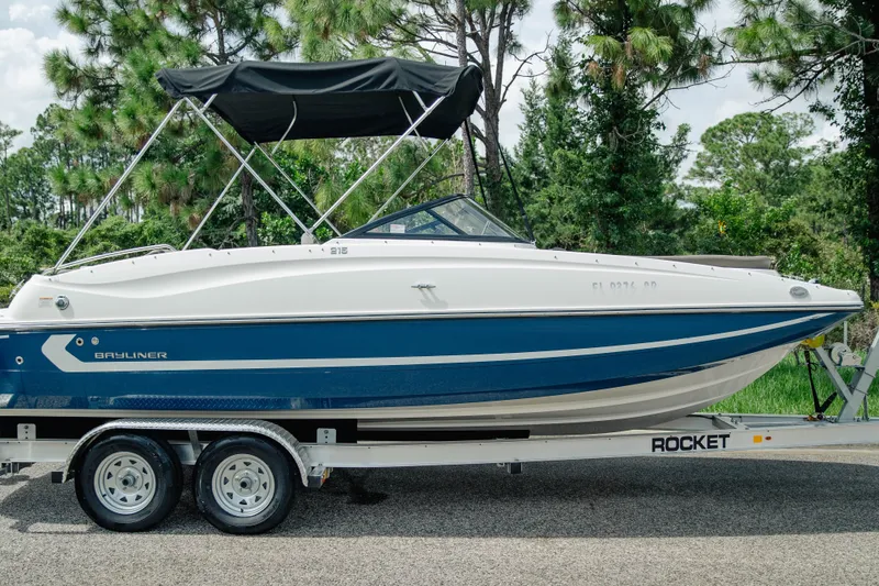 Slide: The Image of 2019 Bayliner 215 Deck Boat on trailer, surrounded by lush greenery. - 0
