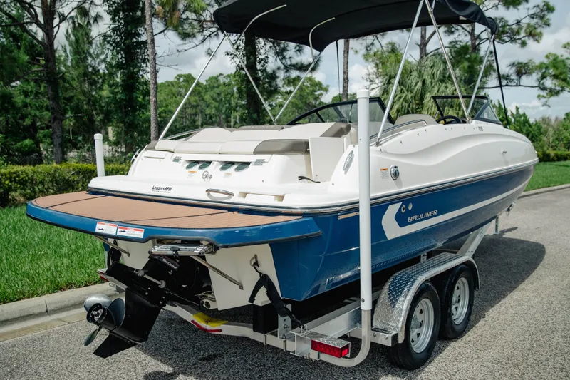 Slide: The Image of 2019 Bayliner 215 Deck Boat on trailer, parked outdoors with lush greenery background. - 6