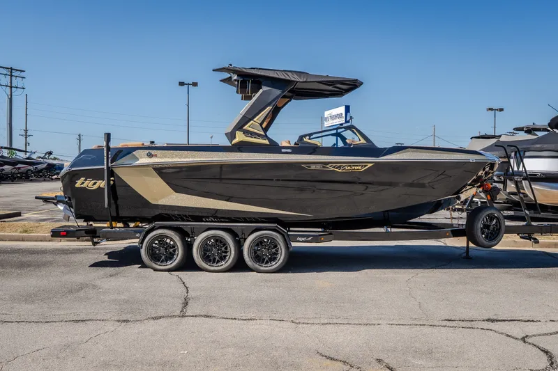 Slide: The Image of 2027 Tigé Ultré 25ZX boat on trailer, sleek black and gold design, parked outdoors. - 2