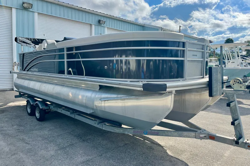 The Image of 2024 Harris Cruiser 230 pontoon boat on trailer, parked outdoors under a partly cloudy sky. - 0