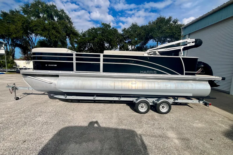 Slide: The Image of 2024 Harris Cruiser 230 pontoon boat on trailer, parked outdoors under a blue sky. - 13