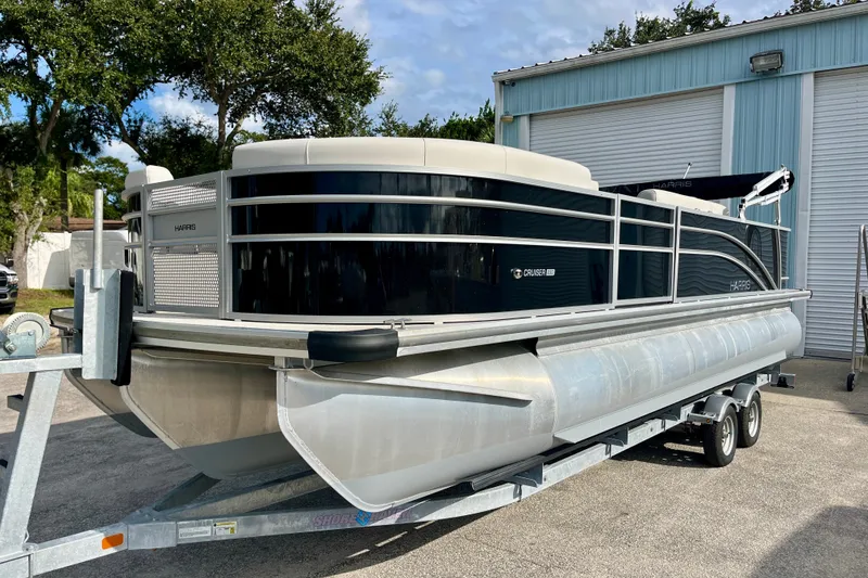 Slide: The Image of 2024 Harris Cruiser 230 pontoon boat on trailer, parked outside a blue building. - 14