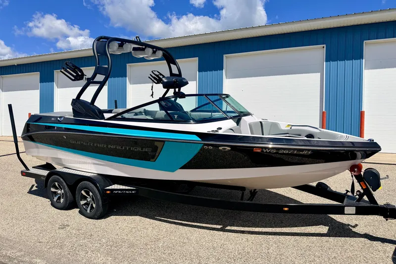 The Image of 2014 Nautique Super Air Nautique 210 boat on trailer, parked outside blue building. - 0