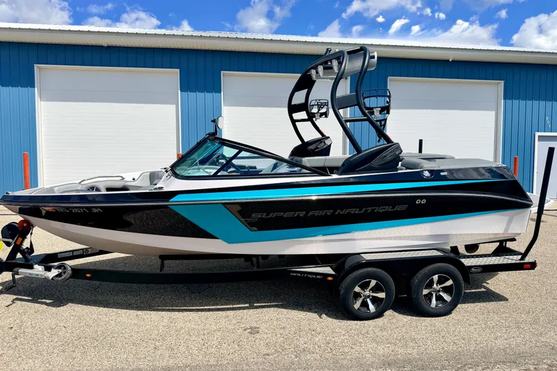 Slide: The Image of 2014 Nautique Super Air Nautique 210 boat on trailer, parked outside blue building. - 1