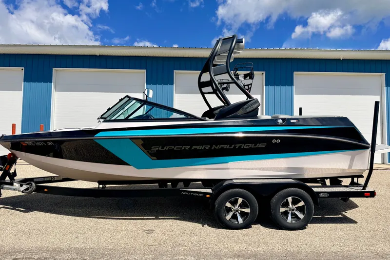 Slide: The Image of 2014 Nautique Super Air Nautique 210 boat on trailer, parked near blue building. - 6