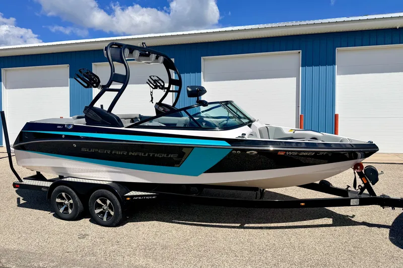 Slide: The Image of 2014 Nautique Super Air Nautique 210 boat on trailer, parked near blue building. - 3