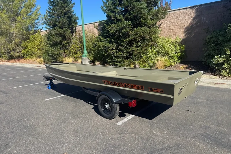 The Image of 2027 Tracker Grizzly 15 Jon boat on trailer in parking lot, surrounded by trees. - 0