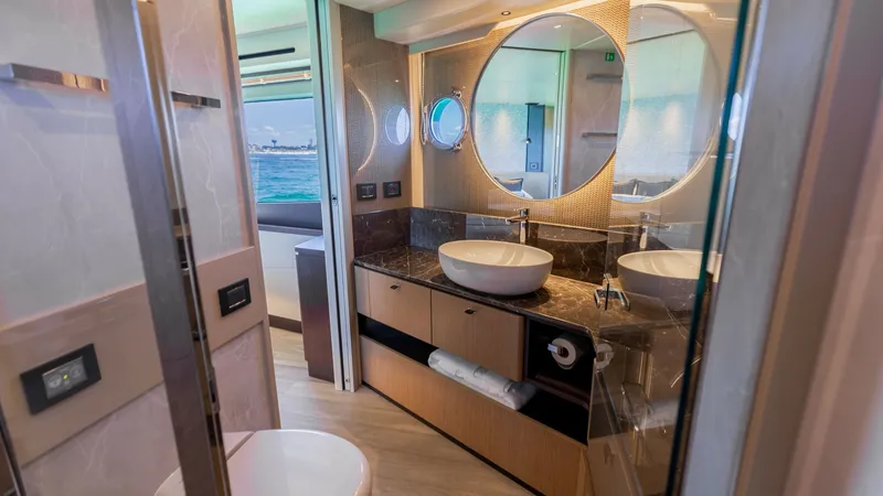 Slide: The Image of Luxurious Master Bathroom This elegant bathroom aboard the 2027 Absolute 60 Flybridge features a modern vanity with a sleek vessel sink and a polished marble countertop. The round mirror and ambient lighting enhance the sense of space and luxury, while the porthole windows offer n - 6492494