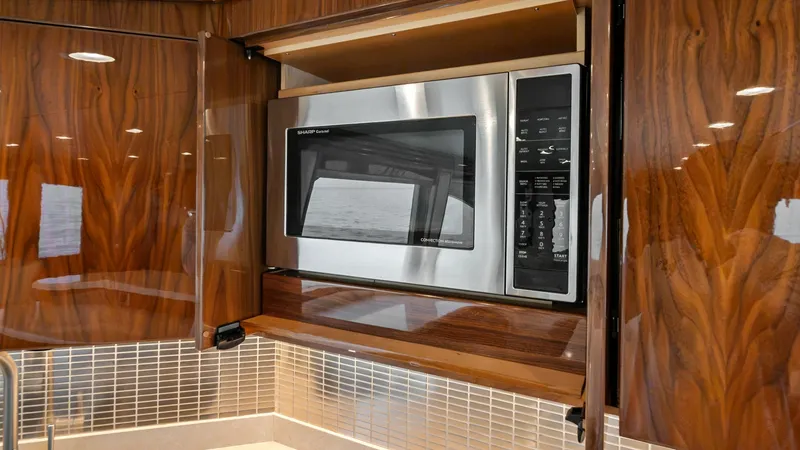 Slide: The Image of Galley Microwave with Wood Cabinetry This image showcases the modern galley of the 2021 Viking 72 Enclosed Bridge, featuring a built-in microwave elegantly integrated into high-gloss wood cabinetry. The sleek design is complemented by a sophisticated tile backsplash and polished countertops, - 7377895