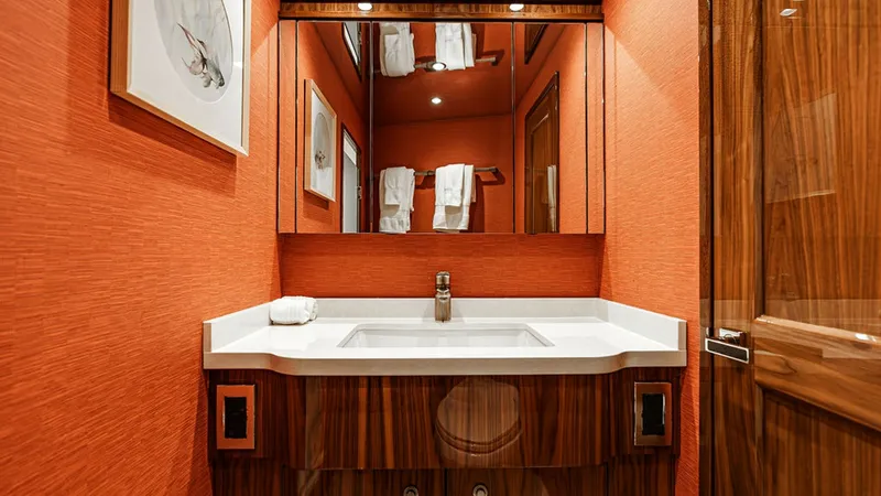 Slide: The Image of Elegant Vanity in Guest Head The guest head of the 2019 Viking 80 Skybridge 'PARTY OF FIVE' features a sophisticated vanity with a sleek, white countertop and integrated sink. Rich wood cabinetry complements the bold, textured orange walls, creating a vibrant and inviting atmosphere. - 6478734