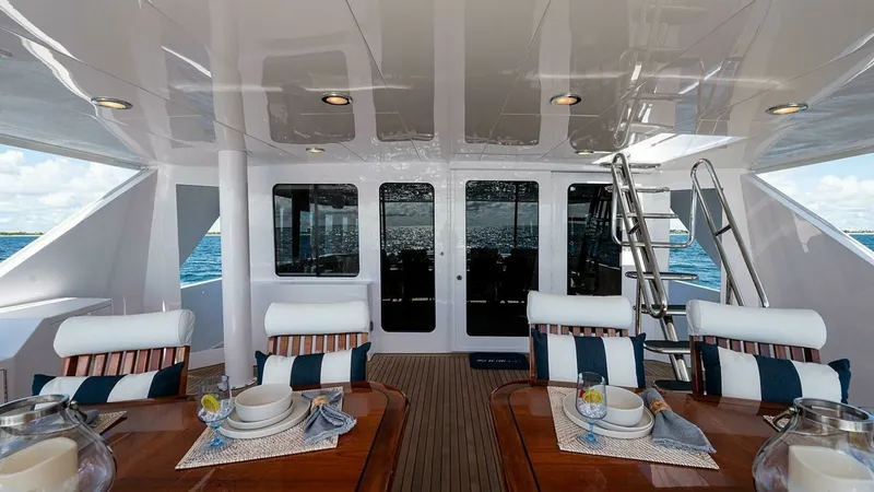 Slide: The Image of Aft Deck with Dining Area The aft deck of the 2003 Hatteras Magnum Ride features an elegant outdoor dining area with two polished wooden tables set for a meal. Comfortable cushioned chairs with striped pillows invite guests to relax and enjoy the ocean views. The area is covered, - 6388435