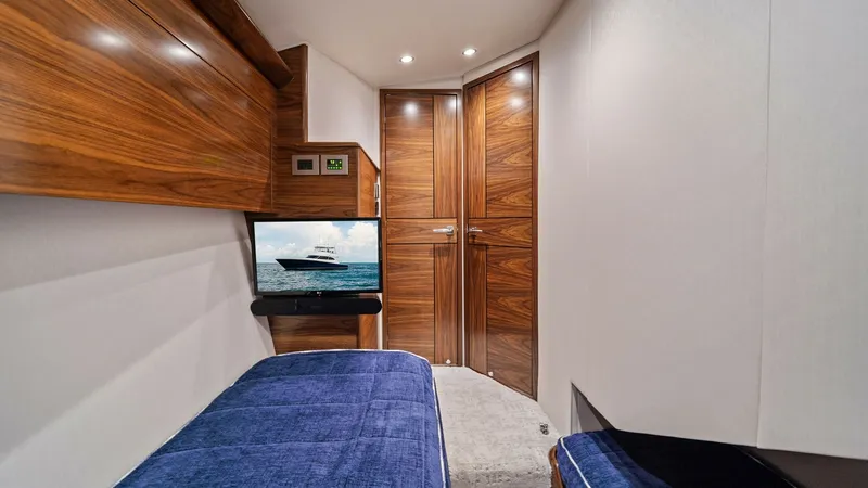 Slide: The Image of Guest Cabin with Twin Berths This cozy guest cabin on the 2024 Viking 64 Convertible features twin berths adorned with plush blue bedding, providing a comfortable retreat for guests. The warm wood finishes and ambient lighting create an inviting atmosphere, while the built-in televis - 7541856