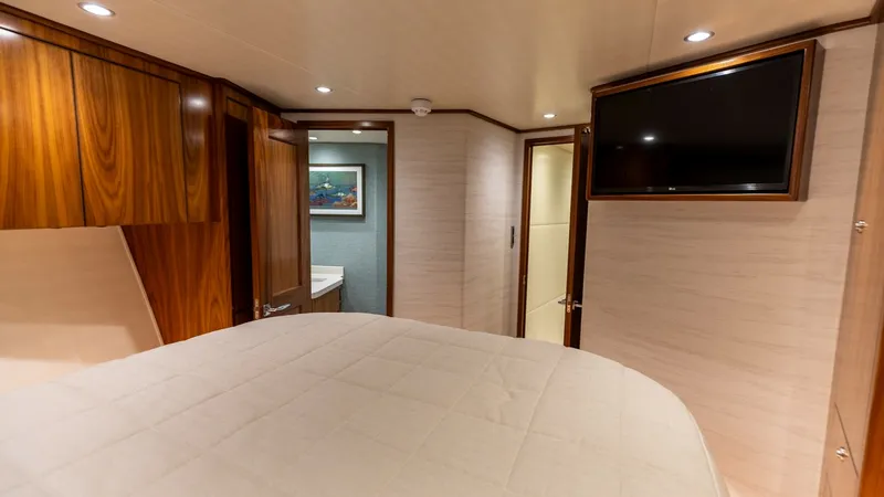 Slide: The Image of Luxurious Forward Stateroom with Ensuite This inviting stateroom on the 2022 Viking 68 Convertible features a plush bed with elegant wooden cabinetry. A flat-screen TV is mounted for entertainment, and the room is softly lit by recessed lighting. The ensuite bathroom is visible through the doorw - 7463814