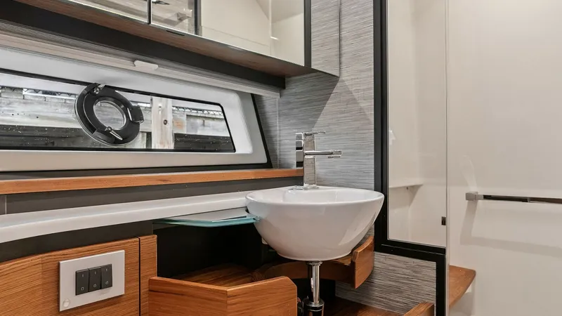 Slide: The Image of Modern Yacht Ensuite Bathroom This elegant ensuite bathroom on the 2025 Tiara 48LE features a sleek, modern design with high-quality materials. The stylish vessel sink is paired with a contemporary faucet, while the wooden cabinetry adds warmth and functionality. A well-placed porthol - 7403647