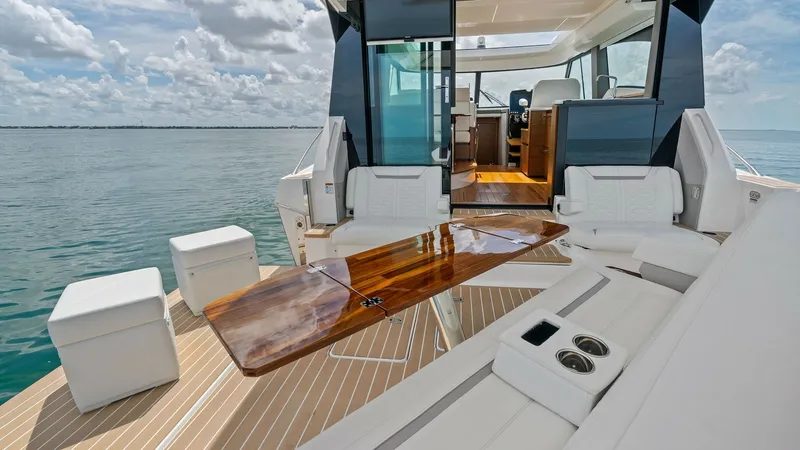 Slide: The Image of Aft Deck with Convertible Seating This spacious aft deck on the 2025 Tiara Yachts 48LE showcases elegant teak flooring and versatile seating arrangements. A sleek, polished wooden table serves as the centerpiece, ideal for alfresco dining or casual gatherings. The open layout seamlessly c - 7403588
