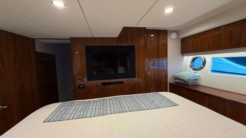 Slide: The Image of Master Stateroom with TV and Portlight The master stateroom of the 2021 Riviera 57 Enclosed Bridge offers a luxurious retreat featuring rich wood paneling and a plush bed. A wall-mounted flat-screen TV provides entertainment, while the portlight allows natural light to enhance the warm interio - 7396146