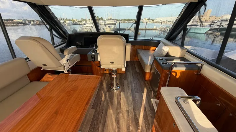 Slide: The Image of Enclosed Bridge Helm and Seating Area The enclosed bridge of the 2021 Riviera 57 offers a commanding helm station with plush captain's chairs and panoramic views. The elegant wood finishes and ample natural light create a welcoming atmosphere. A spacious seating area and well-crafted table pr - 7396135