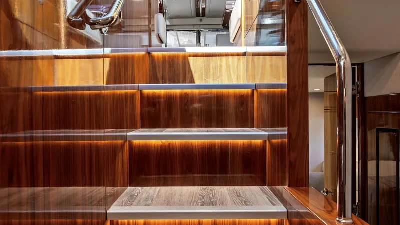 Slide: The Image of Luxury Interior Staircase This image showcases the elegant staircase of the 2021 Riviera 57 Enclosed Bridge. The rich wood finish and sleek stainless steel handrails exude luxury and sophistication. Soft LED lighting highlights each step, creating a warm and inviting ambiance as y - 7541504