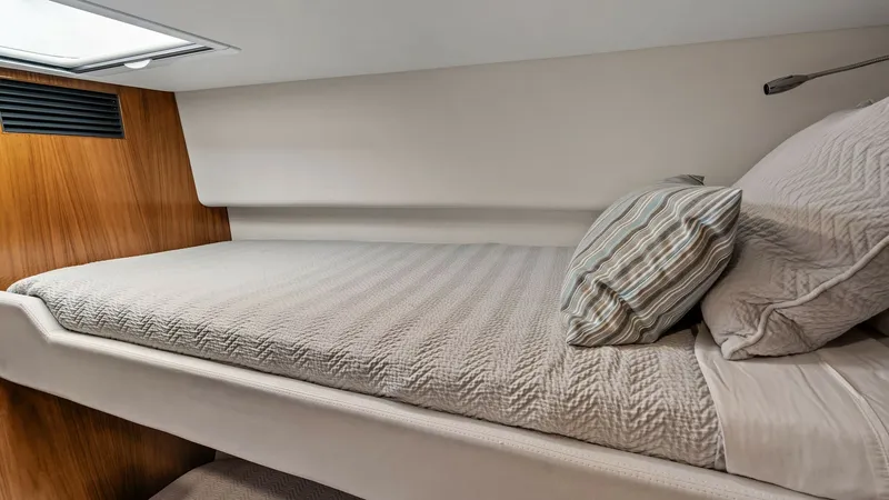 Slide: The Image of Guest Stateroom with Single Berth This cozy guest stateroom on the 2021 Riviera 57 Enclosed Bridge features a comfortable single berth adorned with elegant bedding. The space is accented with warm wood finishes and overhead lighting, creating a welcoming atmosphere. A soft, neutral color - 7541515