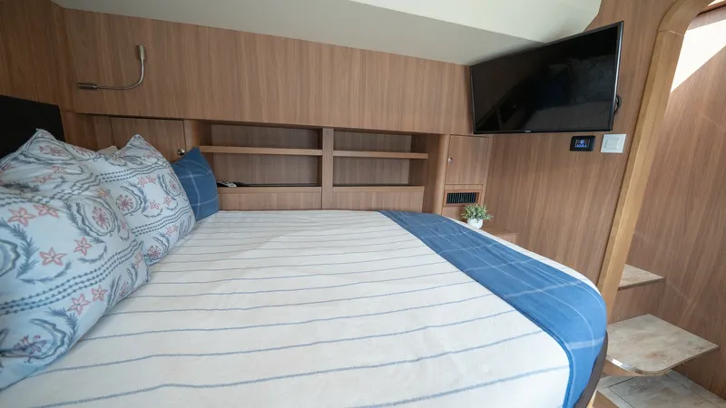 Slide: The Image of Guest Cabin with TV and Storage This inviting guest cabin on the Aquila 44 CAT offers a cozy retreat with a comfortable double bed adorned with nautical-themed linens. The warm wood finishes create a welcoming ambiance, complemented by built-in shelving for ample storage. A wall-mounted - 7413964
