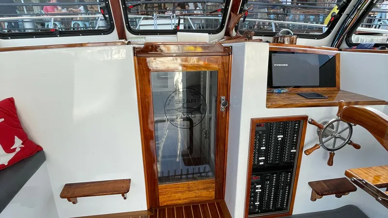 Slide: The Image of Helm Station with Electronics Console This image showcases the helm station of the BEAR FLAG III, featuring a beautifully crafted wooden wheel and an electronics console with modern navigation equipment. The area is accented by rich wood finishes and a sleek glass door leading to the cabin. A - 7397989