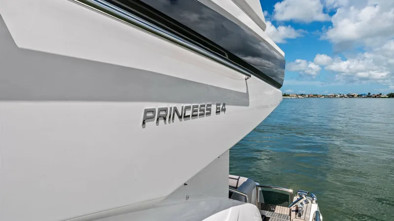 Slide: The Image of Sleek Exterior Profile and Stern Detail This image showcases the elegant exterior profile of the 2016 Princess 64 Flybridge, highlighting its sleek lines and modern design. The vessel's name, 'Princess 64,' is prominently displayed, reflecting its prestigious brand. The view also captures the i - 7460869