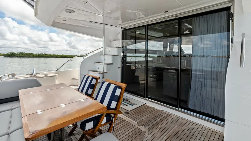 Slide: The Image of Aft Deck with Dining Area This inviting aft deck of the 2016 Princess 64 Flybridge features a sleek teak flooring with a stylish wooden dining table and comfortable seating. The space is elegantly framed by sliding glass doors that lead into the interior, offering a seamless indoo - 7460807