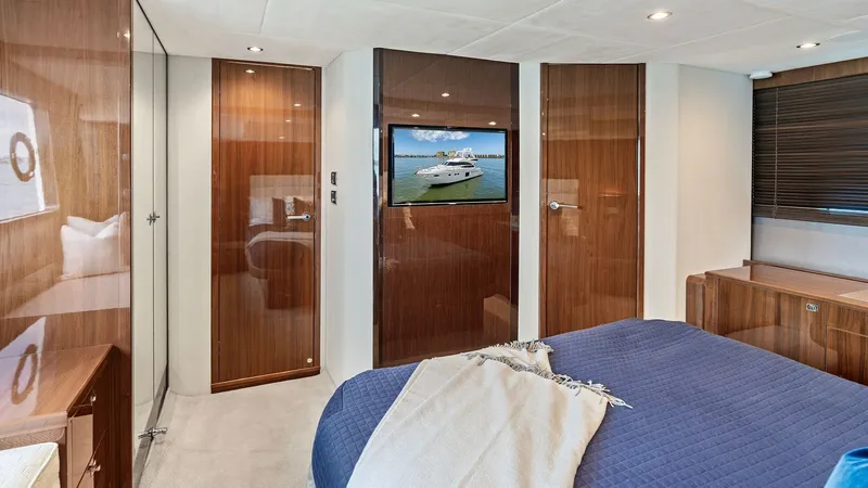 Slide: The Image of Master Stateroom with TV The master stateroom on the 2016 Princess 64 Flybridge features luxurious wood paneling and a plush bed adorned with a soft blue quilt. A flat-screen TV is mounted on the wall, offering entertainment in a comfortable setting. Ample natural light filters t - 7460827