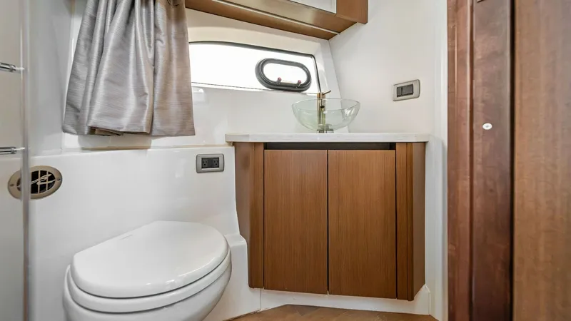 Slide: The Image of Elegant Yacht Bathroom This well-appointed yacht bathroom features sleek wood cabinetry and a modern glass vessel sink, offering both style and functionality. Natural light pours in through the porthole window, complementing the warm wood tones and creating an inviting atmosphe - 7762702