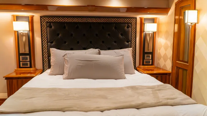 Slide: The Image of Luxurious Master Stateroom Interior This master stateroom exudes elegance with its plush king-sized bed featuring a tufted leather headboard. Warm wood finishes and ambient lighting create a cozy, inviting atmosphere, complemented by stylish bedside lamps and cabinetry. The sophisticated de - 7943370