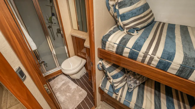 Slide: The Image of Guest Cabin with Ensuite Bathroom This inviting guest cabin aboard the 2019 Viking 68 Enclosed Bridge features cozy bunk beds with striped bedding, perfect for overnight comfort. The ensuite bathroom is compact yet functional, offering a shower, toilet, and stylish wood finishes that enha - 8738030