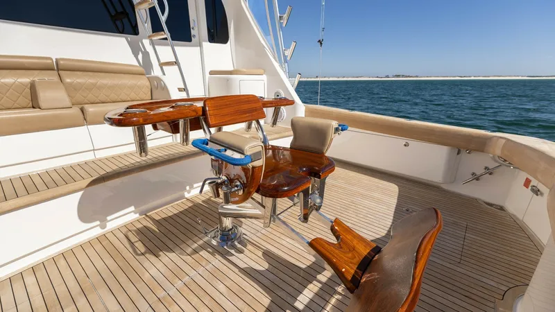 Slide: The Image of Teak Fishing Cockpit with Fighting Chair This image showcases the elegant teak fishing cockpit of the 2019 Viking 68 Convertible. The centerpiece is a robust fighting chair, designed for serious anglers, complemented by a polished wooden table with multiple rod holders. The spacious seating area - 8679296