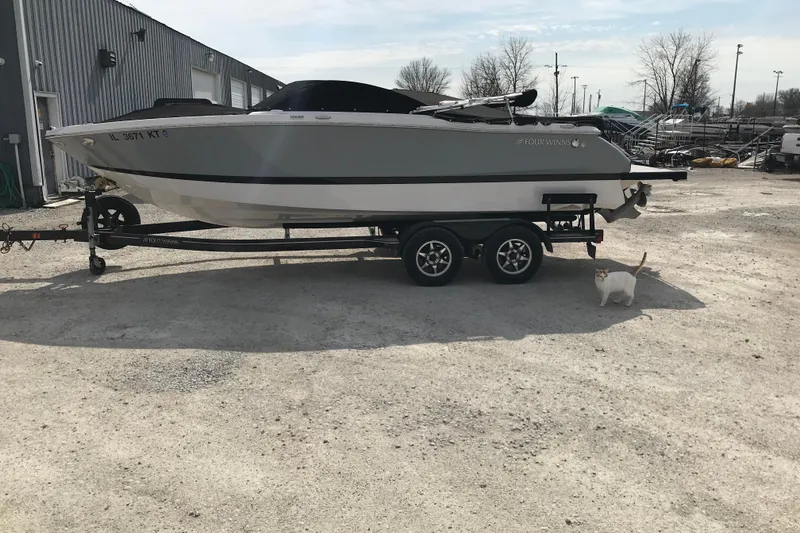 Slide: The Image of 2015 Four Winns H230 boat on trailer with a cat nearby in a gravel lot. - 2