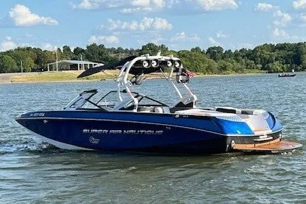 Slide: The Image of A blue 2015 Super Air Nautique G25 boat on a calm lake with a wooded shoreline. - 2