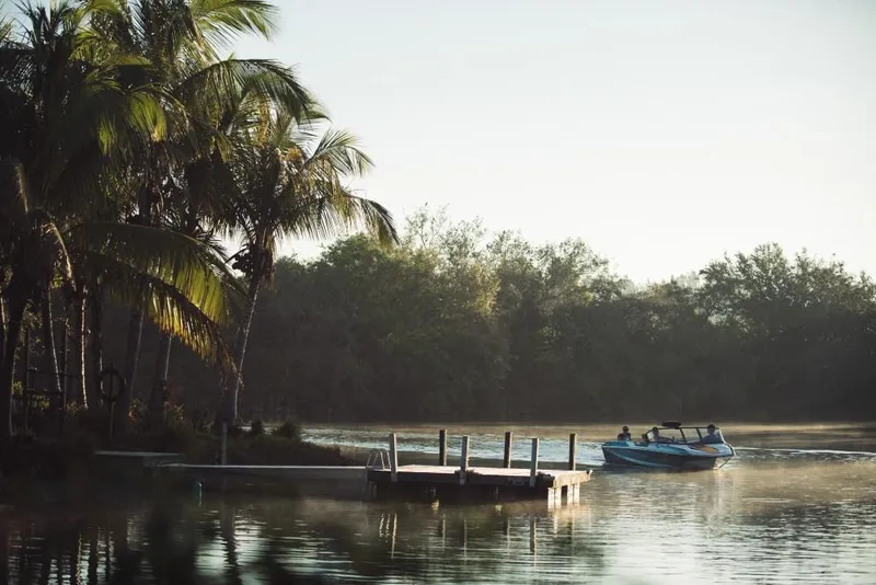 Slide: The Image of 2023 Nautique Ski boat cruising near a dock in a serene, tropical setting. - 10