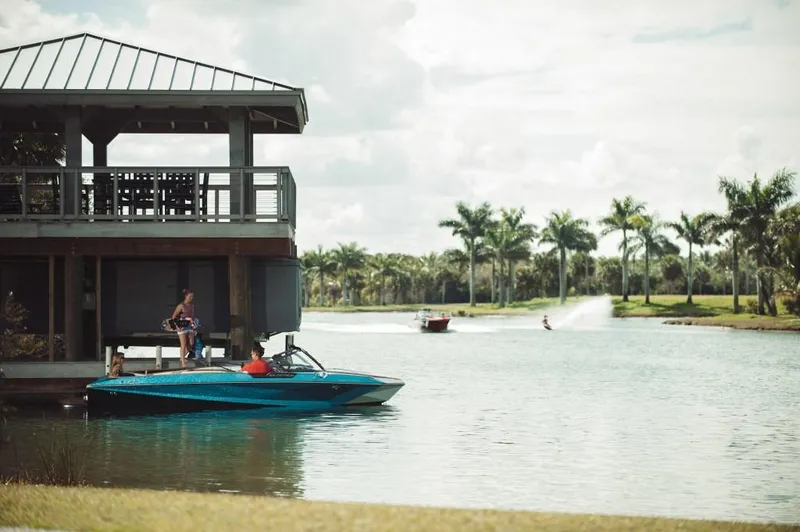 Slide: The Image of 2023 Nautique Ski boat docked by a lakeside house with palm trees. - 12