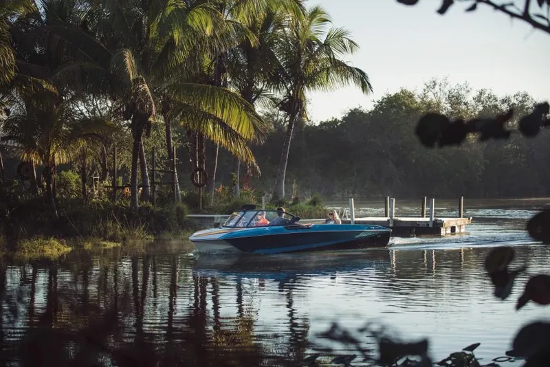 Slide: The Image of 2023 Nautique Ski boat cruising on a serene lake near a dock and palm trees. - 20