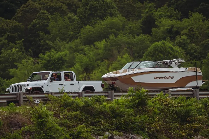 Slide: The Image of White truck towing 2023 MasterCraft XT20 boat on a scenic road with lush greenery. - 3