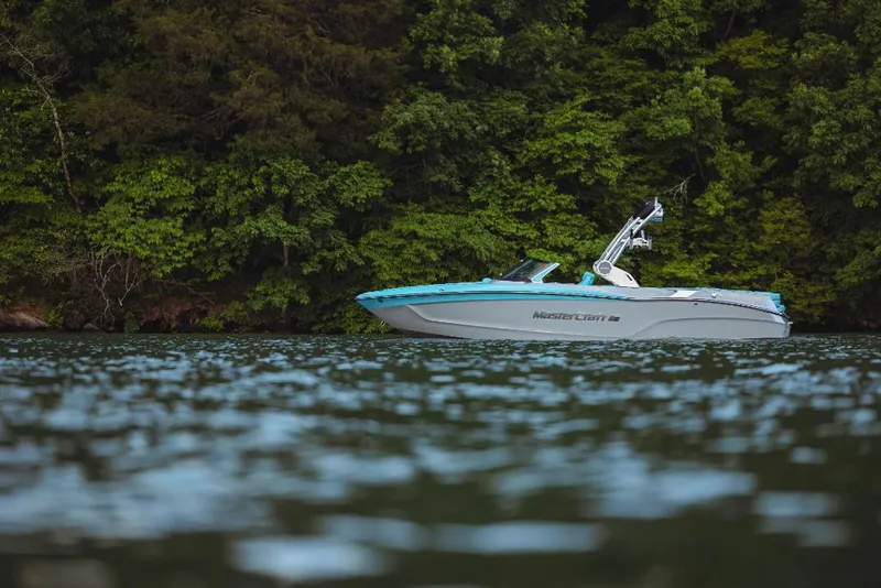 Slide: The Image of 2023 MasterCraft XT23 boat on a serene lake with lush green forest background. - 0