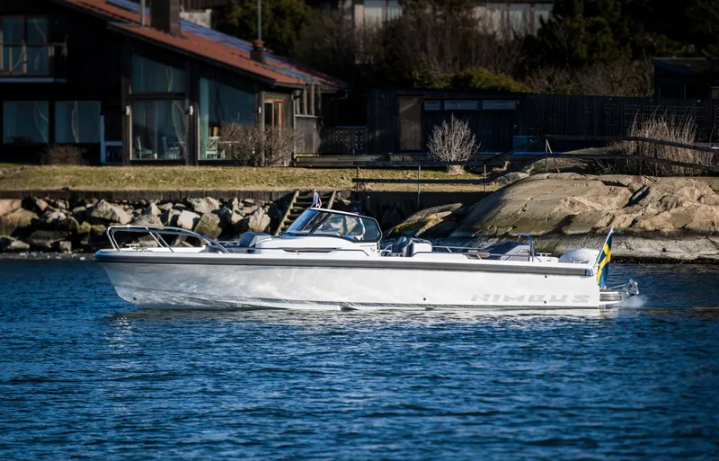 Slide: The Image of Nimbus T9 2025 motorboat cruising near a rocky shoreline with houses in the background. - 0