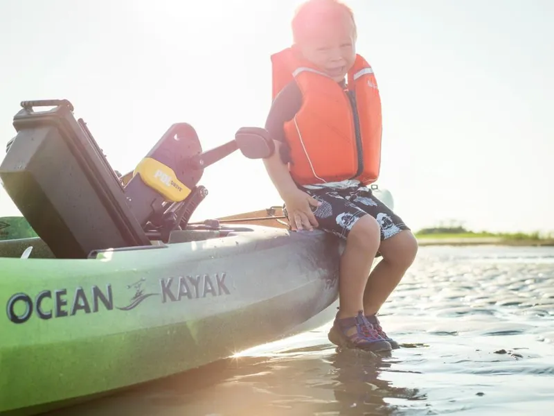 Slide: The Image of Child sitting on 2023 Ocean Kayak Malibu PDL in shallow water, wearing an orange life jacket. - 2