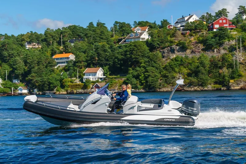 The Image of 2024 Grand Inflatables G850 boat cruising on a scenic lake with houses in the background. - 0