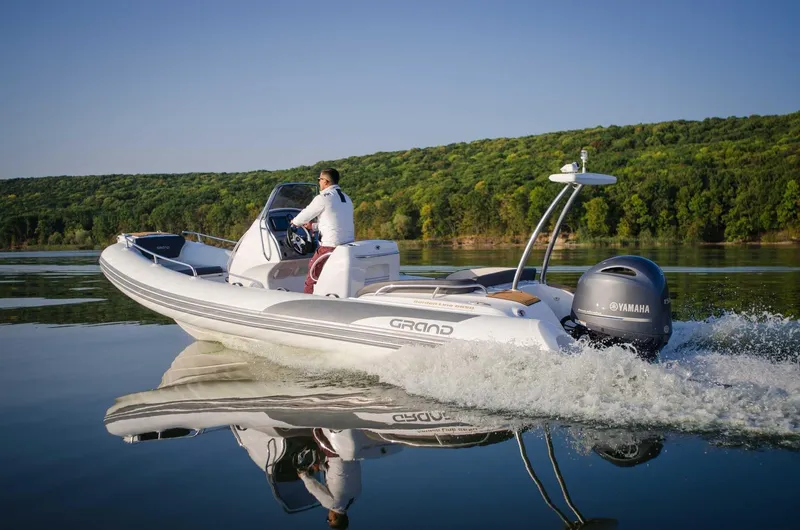 Slide: The Image of 2024 Grand Inflatables G650 boat cruising on a calm lake with lush green hills in the background. - 22