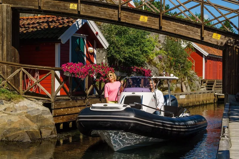 Slide: The Image of 2024 Grand Inflatables G650 boat cruising under a wooden bridge near red cottages. - 5