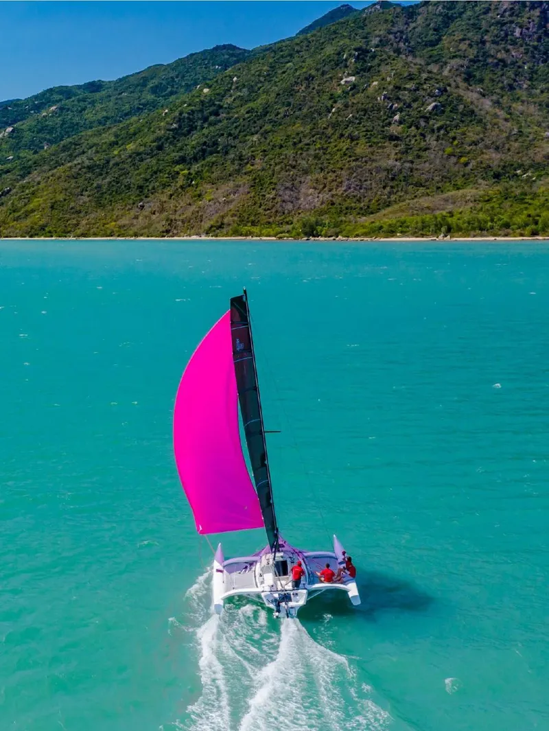 Slide: The Image of Sailboat Corsair 880 (2025) with pink sail on turquoise water near lush coastline. - 2