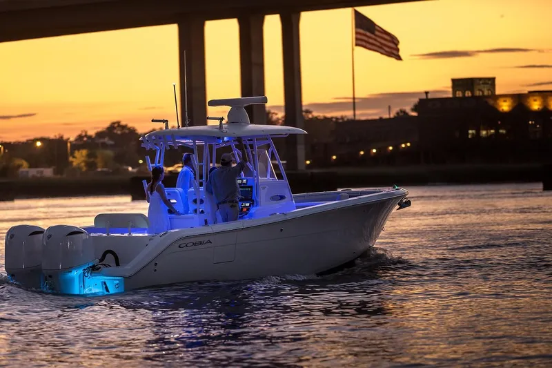 Slide: The Image of 2024 Cobia 301 CC boat cruising at sunset with blue deck lights. - 13