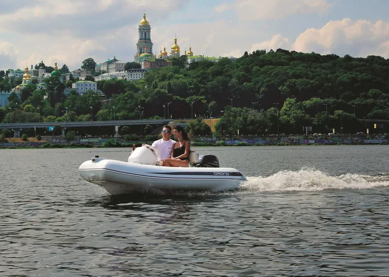 Slide: The Image of 2024 Grand Inflatables S330 boat cruising on a scenic river with historic buildings in the background. - 8