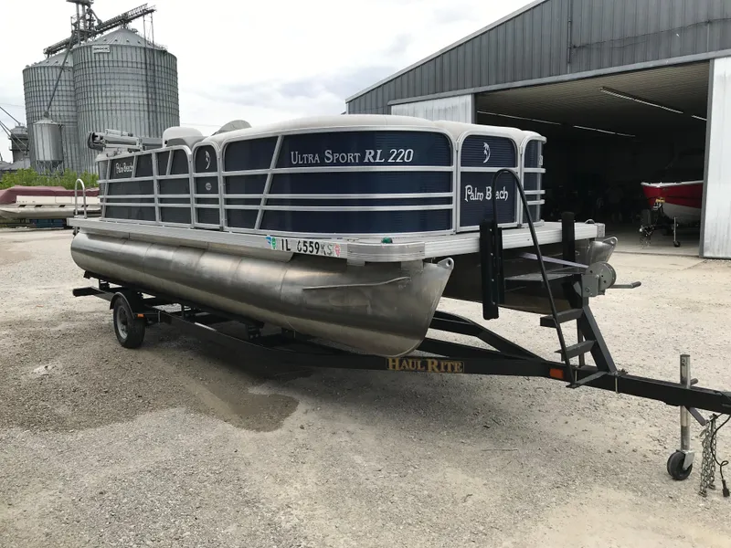 Slide: The Image of 2015 Palm Beach Ultra Sport 220 pontoon boat on trailer, parked near industrial buildings. - 2