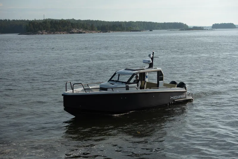 Slide: The Image of 2023 XO Boats Defender 9 cruising on a calm lake near a forested shoreline. - 8